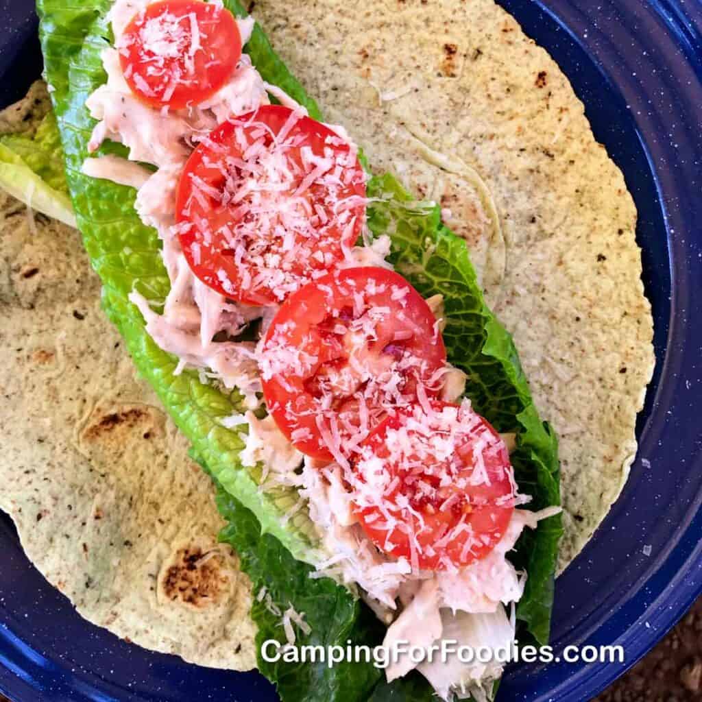 Chicken Parmesan Wrap by CampingForFoodies features a blue camping plate topped with a green spinach tortilla, bright green leaf lettuce, shredded chicken tossed in a ranch salad dressing then finished off with bright red sliced tomatoes and freshly grated Parmesan cheese.