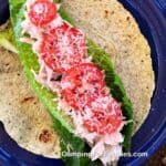 Chicken Parmesan Wrap by CampingForFoodies features a blue camping plate topped with a green spinach tortilla, bright green leaf lettuce, shredded chicken tossed in a ranch salad dressing then finished off with bright red sliced tomatoes and freshly grated Parmesan cheese.