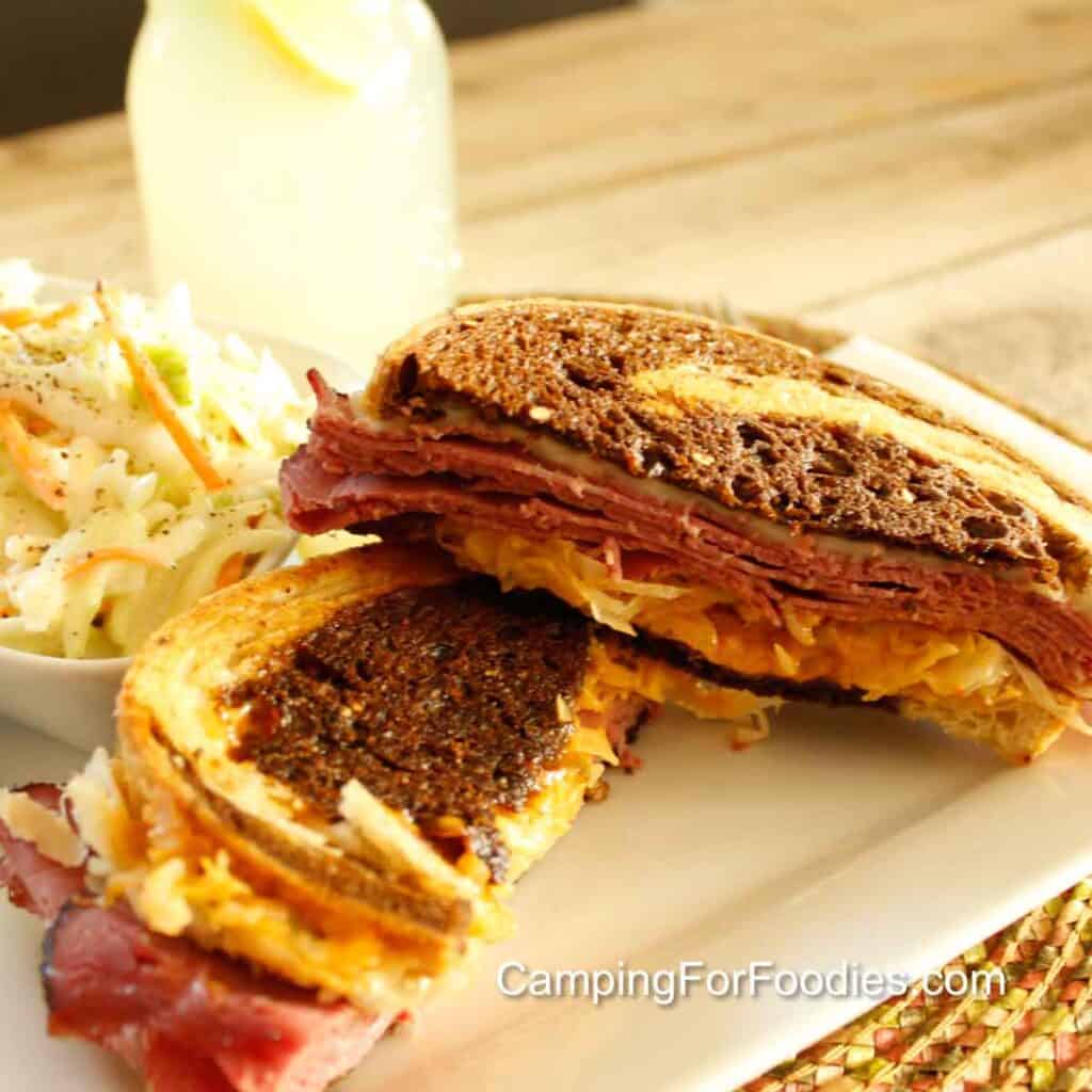 Pastrami Reuben Sandwich by CampingForFoodies features a toasted sandwich cut in half and stacked on a white plate set on a worn picnic table. The brown and golden white swirls in the marble rye grilled bread are stuffed with layers of reddish, pink pastrami deli meat, white Swiss melted cheese, creamy Thousand Island dressing and crunchy sauerkraut. Next to the sandwich is a small bowl of coleslaw, a glass of fresh squeezed lemonade with a lemon slice is in the background.