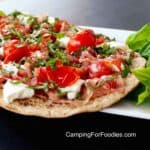 Caprese Flatbread With Prosciutto by CampingForFoodies features a piece of flatbread that has been toasted over a hot grill and topped with thin slices of chopped Prosciutto, chunks of creamy white mozzarella cheese, charred tomatoes and thin ribbons of fresh basil leaves. The white plate is sitting on a black table outside on a sunny day. Bright green fresh basil leaves garnish the plate.