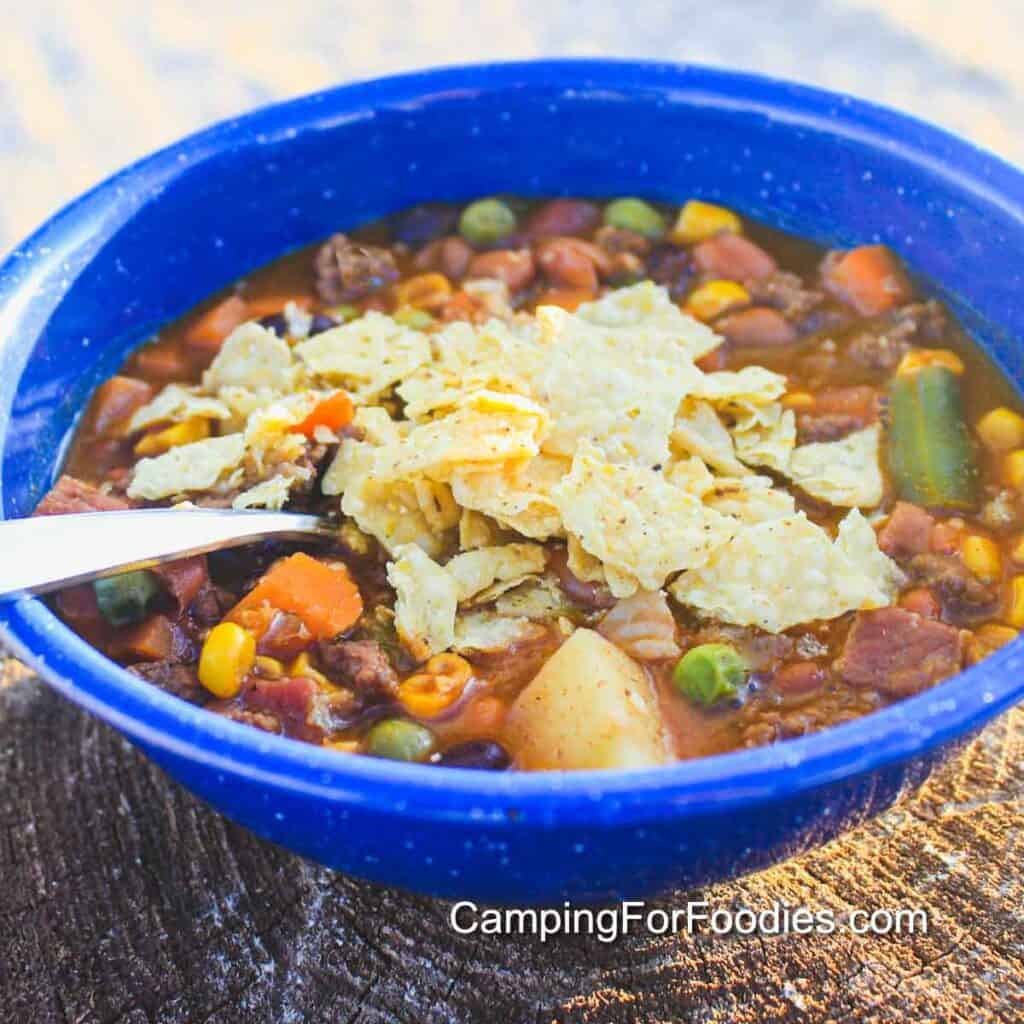 Cowboy Soup Recipe by CampingForFoodies features a blue camping bowl filled with a serving of hamburger Tex-Mex soup in a red tomato sauce broth. Green beans, yellow corn kernels, green peas, orange cubed carrots and white potato chunks in the soup are topped with crushed corn tortilla chips. The handle of a spoon rests on the side of the bowl that is set on a campfire wood log.