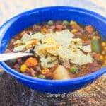 Cowboy Soup Recipe by CampingForFoodies features a blue camping bowl filled with a serving of hamburger Tex-Mex soup in a red tomato sauce broth. Green beans, yellow corn kernels, green peas, orange cubed carrots and white potato chunks in the soup are topped with crushed corn tortilla chips. The handle of a spoon rests on the side of the bowl that is set on a campfire wood log.