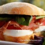 Prosciutto Sandwich Recipe With Fresh Basil And Mozzarella by CampingForFoodies features a ciabatta bread roll sliced in half and filled with bright green fresh basil leaves, pinkish, red colored thinly sliced prosciutto meat, bright red tomato slices and white creamy slices of fresh mozzarella cheese. The sandwich is being served on a white plate in an outdoor setting on a sunny day.