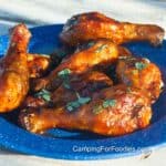 Smoked Honey Sriracha Chicken Drumsticks by CampingForFoodies features a blue camping plate filled with sticky, golden brown, chicken legs coated in brownish, red BBQ sauce that is glistening from the caramelized sugars, bright green pieces of cilantro are scattered across the glazed meat.