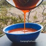 Sriracha BBQ Sauce Recipe by CampingForFoodies features a blue camping bowl being filled with an orangish-red colored sauce. A wire whisk is scraping the pan as the sauce is being poured in a steady stream into the bowl below. The background is a desert setting with desert plants and a very distant mountain range on an overcast day.