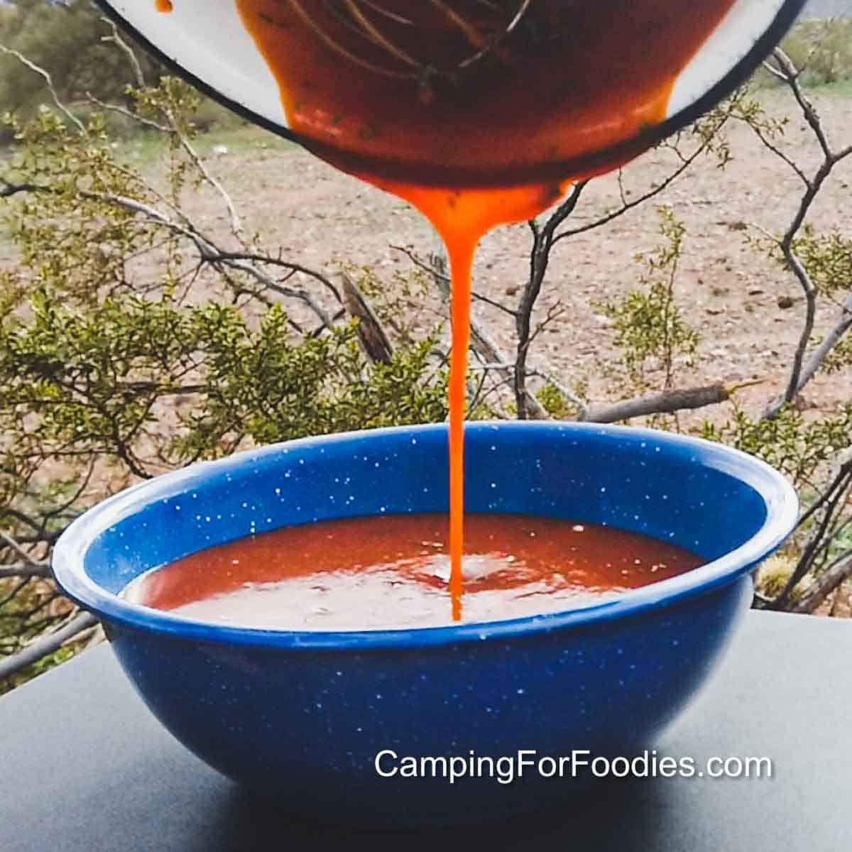 Sriracha BBQ Sauce Recipe by CampingForFoodies features a blue camping bowl being filled with an orangish-red colored sauce. A wire whisk is scraping the pan as the sauce is being poured in a steady stream into the bowl below. The background is a desert setting with desert plants and a very distant mountain range on an overcast day.