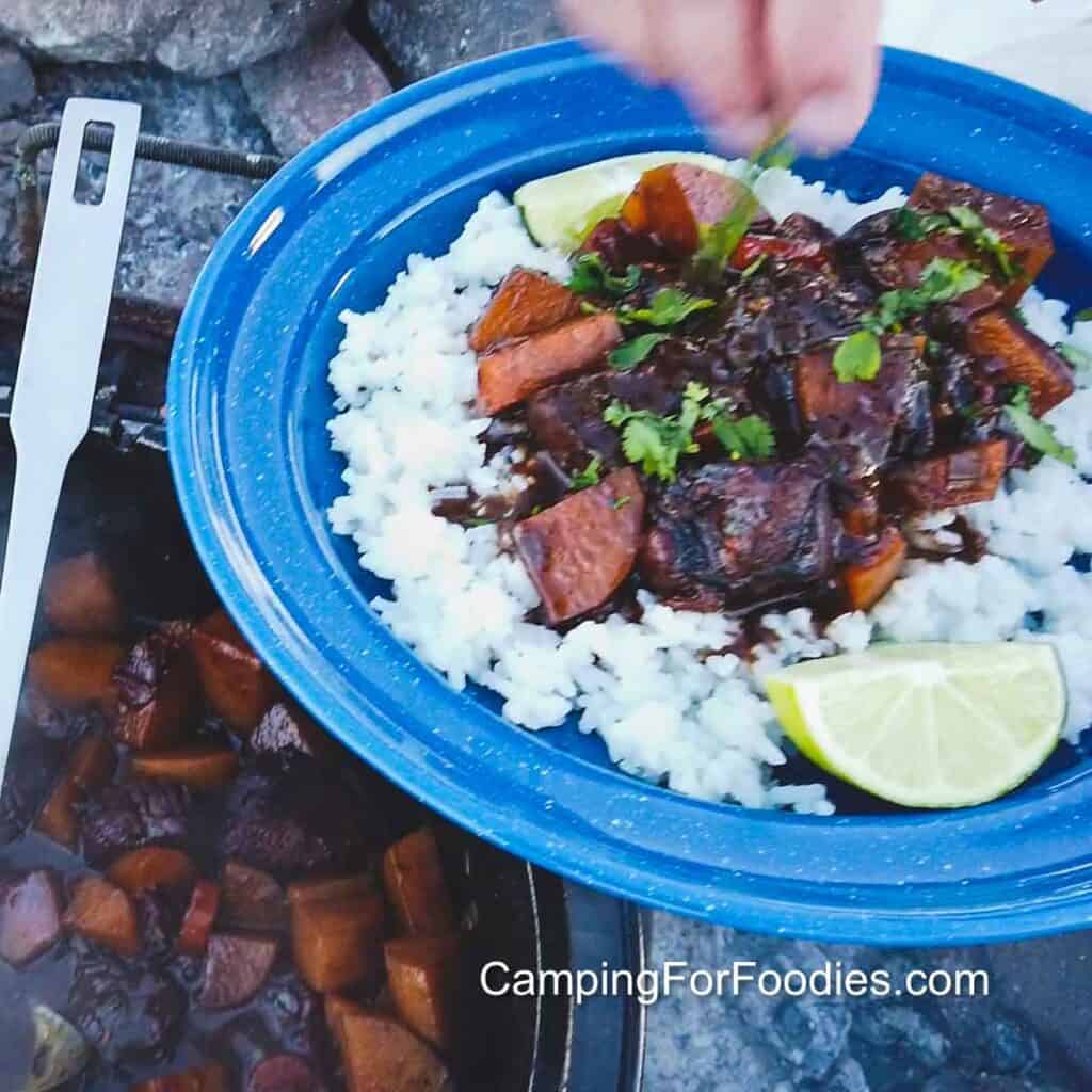 Trinidad Stew Chicken Recipe by CampingForFoodies features a blue camping plate filled with white rice that is topped with deep brown stewed chicken, carrots and potatoes in a thick sauce. A green lime wedge rests on the side while a hand sprinkles bright green cilantro over the plate. A Dutch oven in the background contains more stewed chicken with the handle of a ladle resting on the edge of the pot.