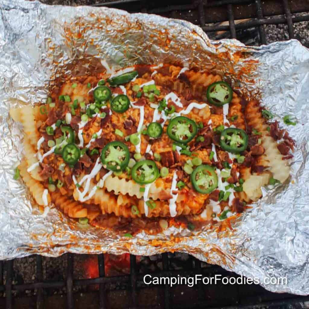 Campfire Chili Cheese Fries by CampingForFoodies features a shiny silver colored piece of tin foil sitting on a campfire grate with orange glowing coals in the background. The crinkle cut French fries are topped with steaming hot chili, melted cheddar cheese, creamy white drizzles of sour cream and sliced green onions and jalapeno peppers.