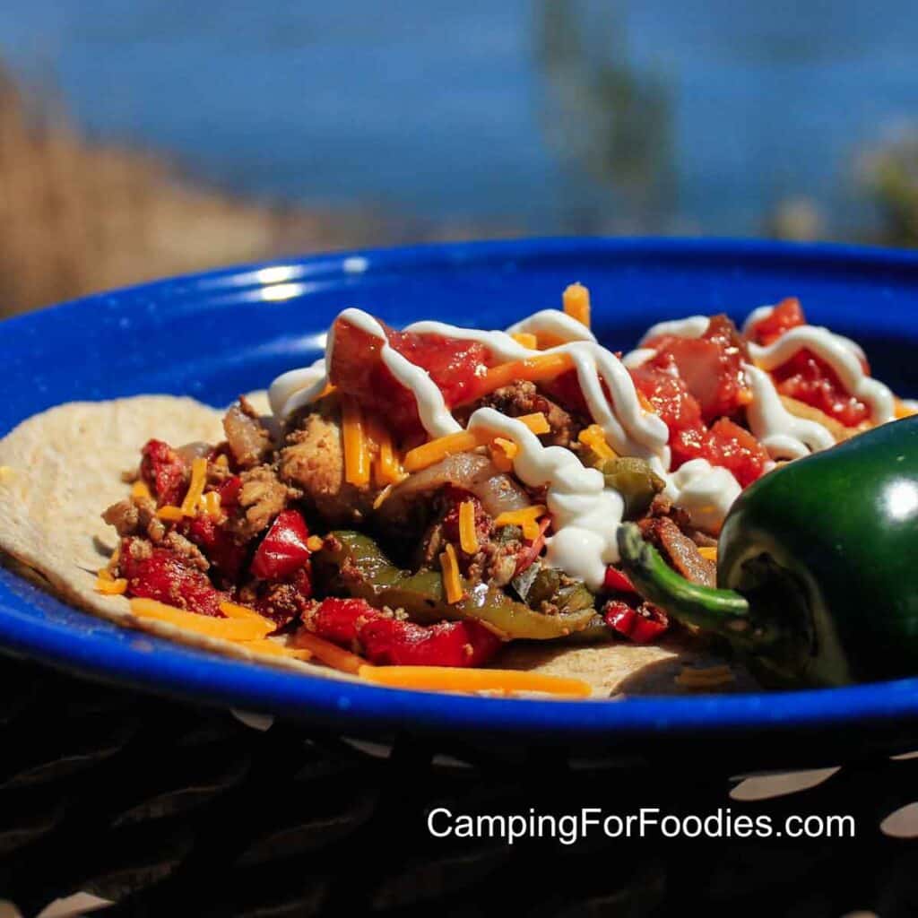 Chicken Fajitas Freezer Meal by CampingForFoodies features a blue camping plate holding a brown, whole-wheat tortilla topped with golden brown chicken fajitas, red and green peppers, shredded cheddar cheese, spicy red salsa and drizzles of white, creamy sour cream. A green jalapeno pepper sits at the side of the plate on a picnic table with a blue, flowing river in the background on a bright sunny day.