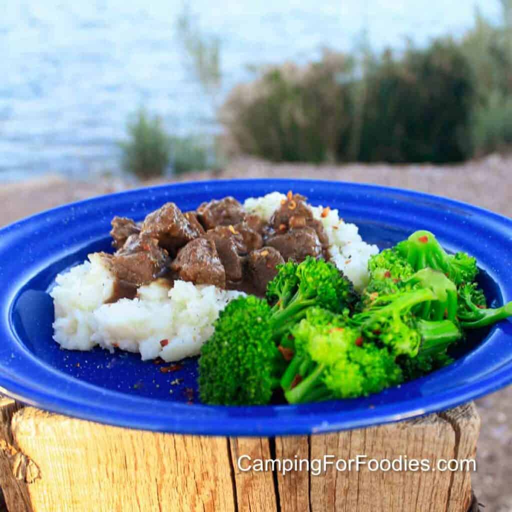 Kettle Beef Recipe by CampingForFoodies features a blue camping plate filled with fluffy, white mashed potatoes topped with tender beef chunks in a thick brown gravy sauce alongside bright green steamed broccoli. Both are sprinkled with red pepper flakes. The plate is set on a campfire log. In the background is a sparkling river and tall green grasses.