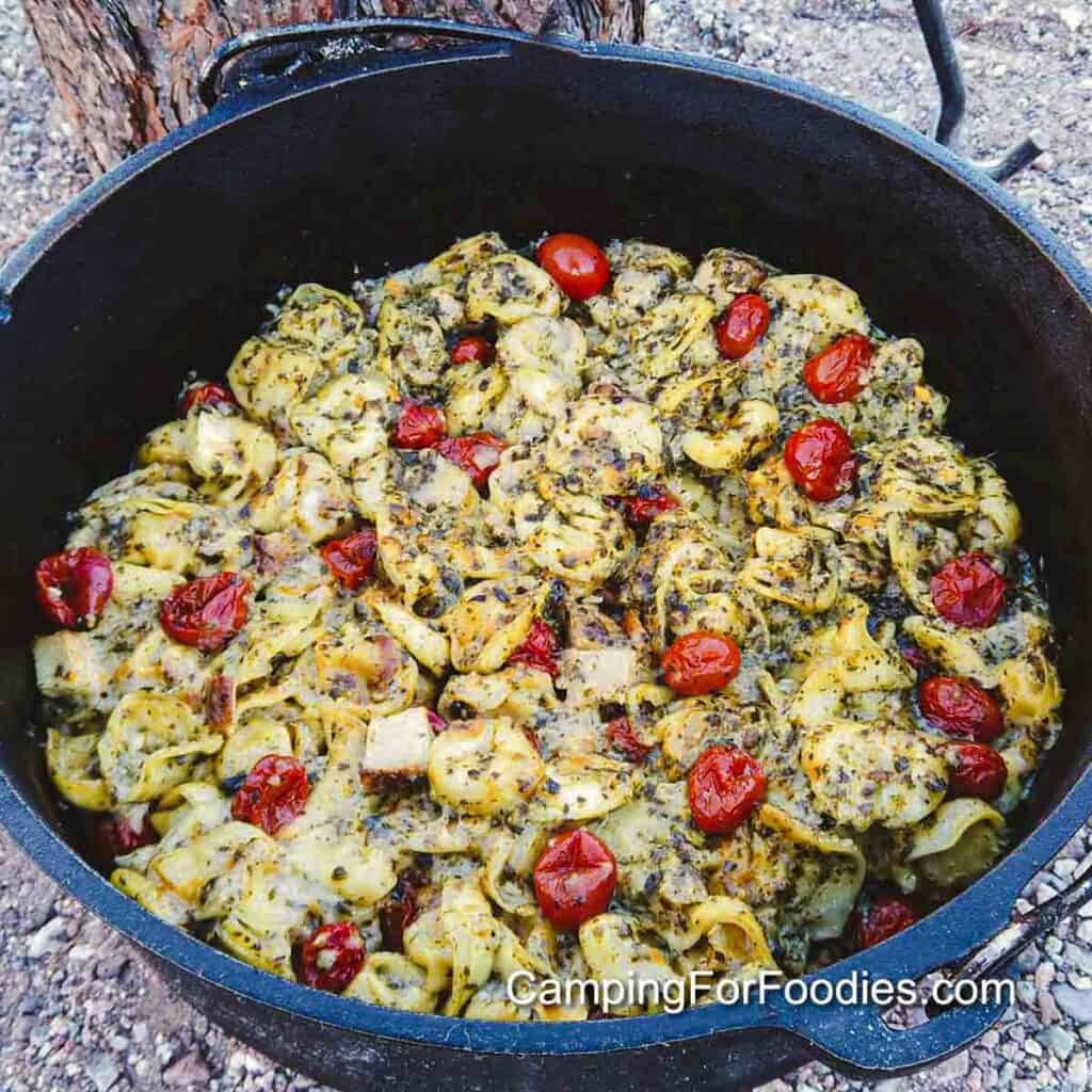 Pesto Tortellini Bake Freezer Meal by CampingForFoodies features a black cast iron Dutch oven filled with cubed chicken, red cherry tomatoes and cheese-filled tortellini pasta in a creamy pesto and alfredo sauce. The family-style meal has been baked and is ready to be served.