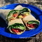Raw Veggie Wraps Recipe by CampingForFoodies features a blue camping plate set on a log outdoors in bright sunshine. It is filled with two raw vegan wraps that are cut in half and stacked on top of each other. The brown wheat tortillas are filled with green chopped spinach, sliced avocados and cucumbers, black beans, red tomatoes, bell peppers and onions, brown sunflower seeds and creamy hummus.