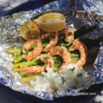 Shrimp And Asparagus Foil Packs by CampingForFoodies features a piece of shiny aluminum foil set on a table outside on a sunny day. The foil pack holds 6 cooked jumbo shrimp set on a stack of bright green asparagus and topped with chopped fresh parsley, on the side is fluffy white rice and two lemon wedges.