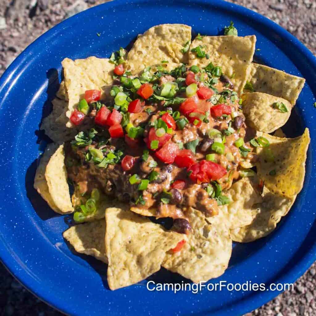 Creamy Taco Recipe by CampingForFoodies features a blue camping plate filled with a serving of creamy beef tacos. Corn tortilla chips are covered with browned ground beef and black beans in a thick brownish-orange cheesy sauce that’s topped with bright red diced tomatoes, sliced green onions and bright green cilantro.