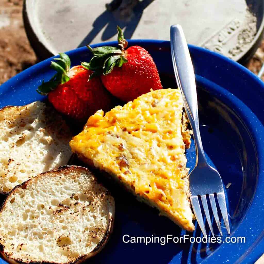 Dutch Oven Egg Bake by CampingForFoodies features a blue camping plate filled with a breakfast meal including a slice of golden brown and yellow egg casserole baked in the black cast iron Dutch oven setting in the background, on the side are two bright red strawberries, toasted English muffins and a shiny fork all glistening from the sunshine on a bright morning.