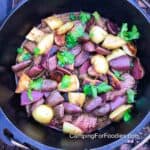 Dutch Oven Steak by CampingForFoodies features a black cast iron camp Dutch oven filled with strips of steak in a sauce with tender white and purple fingerling potatoes. The dish is topped with bright green fresh parsley. The pot is set on top of a grate that is set on the ground outdoors.