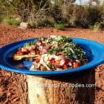 Grilled Flatbread Pizza Recipe by CampingForFoodies features a blue camping plate filled with toasted flat bread that’s topped with bright red cherry tomatoes, creamy white mozzarella cheese, kalamata olives and ribbons of bright green basil. The plate is set on a campfire log on a sunny day with a desert landscape in the background.