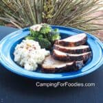Grilled Pork Loin Roast by CampingForFoodies features a blue camping plate filled with 4 slices of grilled pork loin with a golden-brown crust, the meat is set next to mashed potatoes and steamed broccoli that are sprinkled with red pepper flakes. The table is set outside on a sunny day with a desert landscape in the background.
