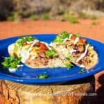 Grilled Steak Burrito Recipe by CampingForFoodies features a blue camping plate filled with a grilled stuffed burrito that is cut in half. The charred, golden brown tortilla is filled with thin strips of steak, black beans, cilantro rice and melted cheese. White sour cream drizzles cover the food along with red salsa and bright green cilantro. The plate is set on a campfire log on a sunny day.