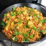 One Pot Camping Cast Iron Fish Recipe by CampingForFoodies features a black cast iron skillet on a camp stove filled with cubed white fish nestled in a bed of brown rice with chopped red tomatoes and bright green parsley leaves.