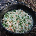 Chicken And Rice Camping Recipe by CampingForFoodies features a black cast iron Dutch oven filled with a baked chicken and rice casserole. The cubed white chicken is smothered in a creamy sauce with white jasmine rice, orange cubed carrots, green sweet peas and bright green fresh parsley. A Dutch oven lid with charcoal briquettes covered in ash sits in the background.