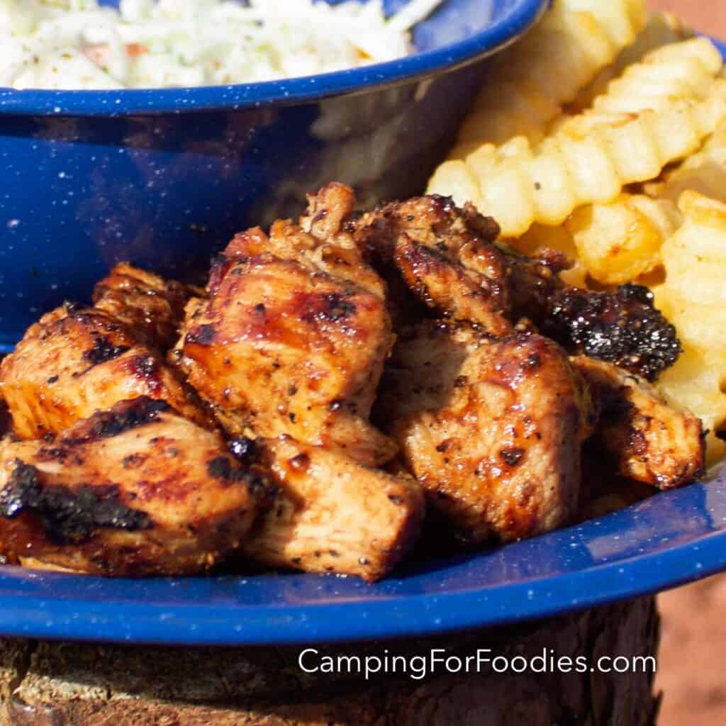 Grilled Chicken Nuggets Recipe by CampingForFoodies features a blue camping plate filled with golden brown chicken nuggets. The char marks and caramelized bbq sauce glisten in the sun. Golden brown French fries sit behind the meat alongside a blue bowl filled with coleslaw in a creamy dressing.