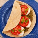 Pesto Tomato Camping Wraps by CampingForFoodies features a blue camping plate filled with a wheat tortilla topped with green basil pesto, fresh sliced red roma tomatoes, creamy white slices of fresh mozzarella cheese and fresh basil leaves. The tortilla is partially folded in preparation for serving.