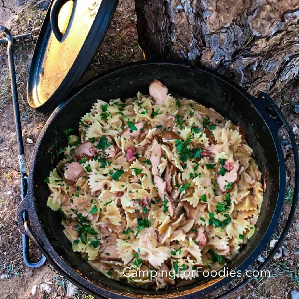 Cajun Chicken One Pot Camping Pasta by CampingForFoodies features a black cast iron Dutch oven filled with chicken and pasta tossed in a creamy Cajun sauce. The pot is set on the ground next to a Dutch oven lid lifter, the lid is resting against a tree in the background.