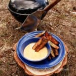 Crispy Grilled Potato Wedges by CampingForFoodies features a blue camping plate filled with charred potato wedges. A blue camping bowl is next to the wedges, it’s filled with a light red colored Chipotle Mayonnaise dipping sauce. The plate is set on a log in front of a small charcoal grill on the ground in the background with a wooden-handled spatula.