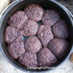 Dutch Oven Chocolate Lava Cakes by CampingForFoodies features a black cast iron camp oven filled with freshly baked individual servings of mini chocolate cakes in foil cupcake wrappers. The pot is set on the ground next to a rock and piece of firewood.