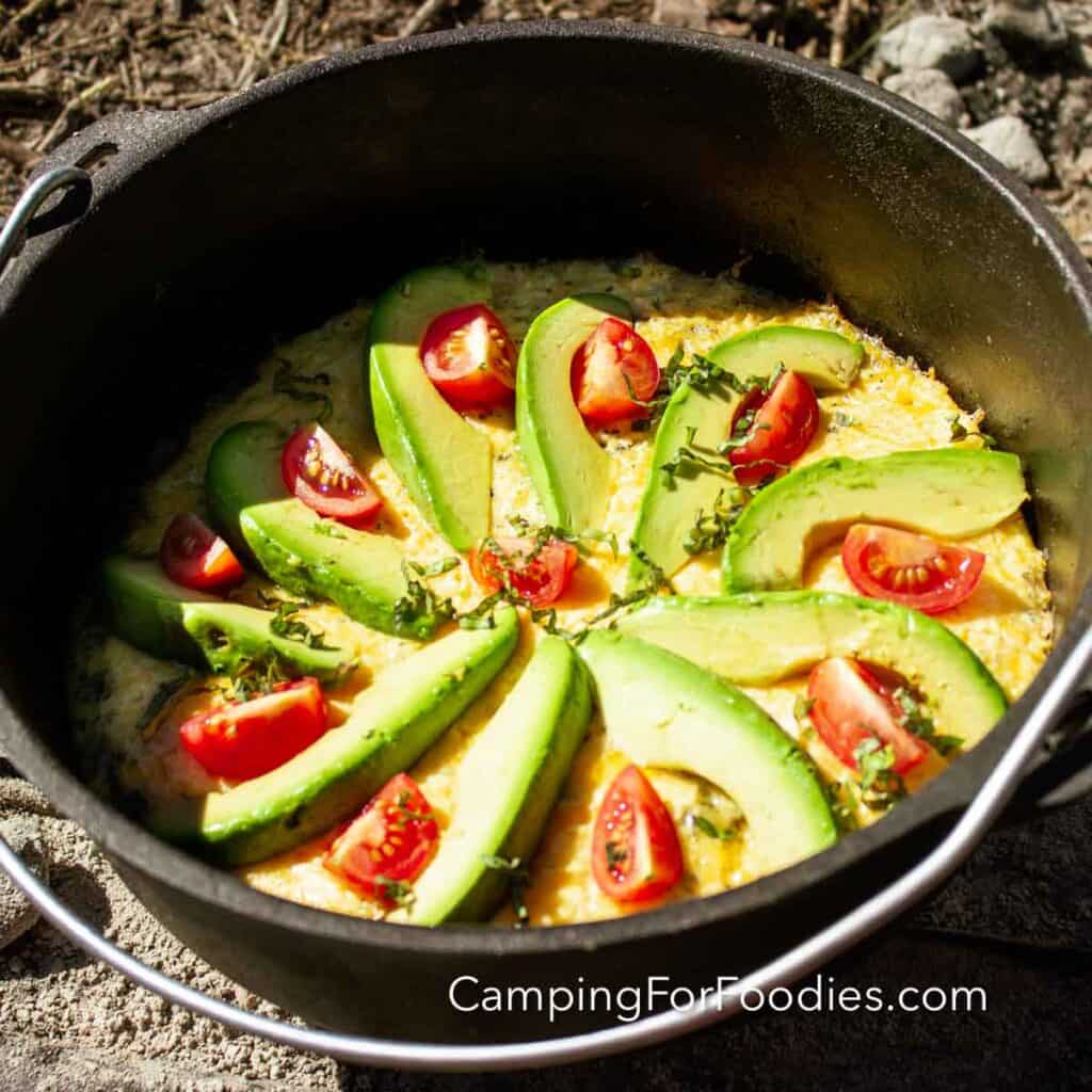 Tomato And Avocado Dutch Oven Frittata by CampingForFoodies features a black cast iron camp oven filled with a freshly baked, golden yellow egg breakfast meal. Bright green slices of avocados and bright red cherry tomatoes top the dish along with ribbons of dark green fresh basil. The pot is set on the ground on a bright sunny day.