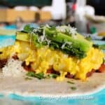California-Style Camping Breakfast Burritos by CampingForFoodies features a white flour tortilla topped with fluffy scrambled eggs and cubed potatoes, reddish-brown chorizo sausage and black beans. The pile is topped with bright green avocado slices, chopped cilantro and ribbons of creamy white shredded Monterey jack cheese. The tortilla is laying flat, waiting to be folded. A camp stove, salt and pepper are in the background on a sunny day.