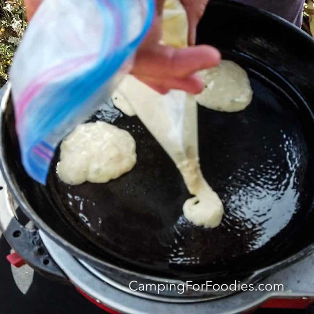 Camping Pancake Mix Plastic Bag Pancakes Hack by CampingForFoodies features a Ziploc bag filled with pancake batter. A hand is holding the bag with a corner snipped off allowing batter to be piped into a black cast iron skillet.