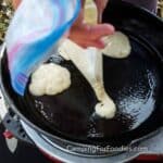 Camping Pancake Mix Plastic Bag Pancakes Hack by CampingForFoodies features a Ziploc bag filled with pancake batter. A hand is holding the bag with a corner snipped off allowing batter to be piped into a black cast iron skillet.