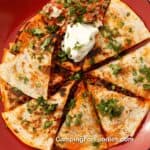 Chorizo Quesadilla by CampingForFoodies features two golden brown crispy tortillas that are sandwiched together and stuffed with a meat, potato and cheese mixture. The toasted quesadilla is sliced into wedges that are set on a red camping plate. A large scoop of red, white and green colored salsa and large dollop of creamy white sour cream is next to the wedges. The entire plate is sprinkled with bright green fresh cilantro which is lit by a setting afternoon sun.