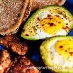 Egg Stuffed Avocados Dutch Oven Recipe by CampingForFoodies features a blue camping plate filled with two eggs baked in an avocado that has been split in half. The creamy yellow egg yolks and egg whites are topped with black pepper and red pepper flakes. Breakfast sausage patties and toasted English muffins are also on the plate that is lit by the morning sun.
