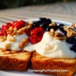 Grilled Pound Cake With Berries by CampingForFoodies features a plate filled with two slices of grilled pound cake that has golden brown grill marks. The slices are topped with a creamy white mascarpone cheese mixture. Red fresh raspberry and fresh blueberry toppings are spooned over the mascarpone mixture and topped with toasted walnuts. The afternoon setting sun is gently lighting the food.