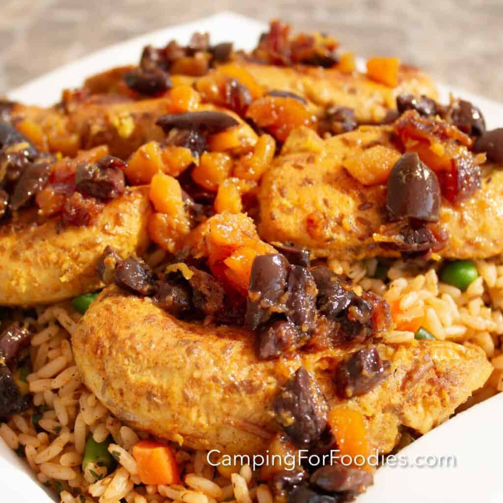 Middle Eastern Chicken by CampingForFoodies features a plate filled with orange and yellow colored chicken strips tossed with orange apricot pieces, brown date pieces and chopped kalamata olives. The dinner is served on a bed of rice and vegetables. The meal is glistening from the sun on a bright afternoon.