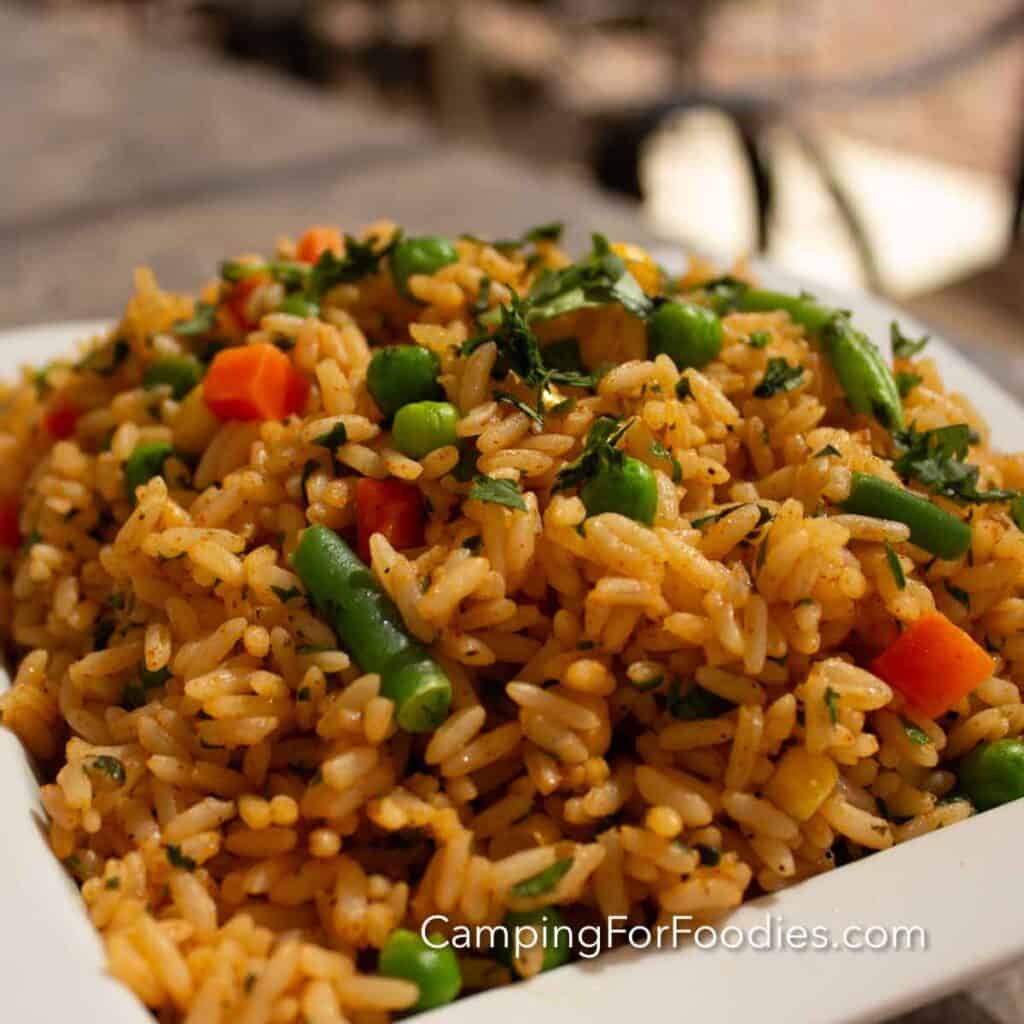 Moroccan Spiced Rice by CampingForFoodies features a large bowl of golden colored rice that is tossed with orange diced carrots, bright green peas and green beans. Fresh green herbs are chopped and tossed with spices to make a flavorful dressing coating the rice and vegetables. The side dish is set on a picnic table as the setting sun dimly lights the food.