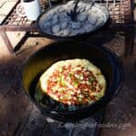 Savory Dutch Baby Camping Recipe by CampingForFoodies features a black cast iron camp oven set on the ground. The pot is filled with a puffy breakfast pancake topped with golden brown crumbled bacon, melted cheese and sliced green onions. Behind the pot is a campfire grate holding the oven’s lid with ash covered charcoal pieces, a cup of coffee is in the background on a bright sunny morning.