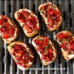 Strawberry Basil Bruschetta by CampingForFoodies features six slices of toasted golden-brown crostinis topped with creamy white goat cheese and macerated strawberries in a balsamic glaze and ribbons of bright green basil. They are set on a black grill grate of a propane camp grill.