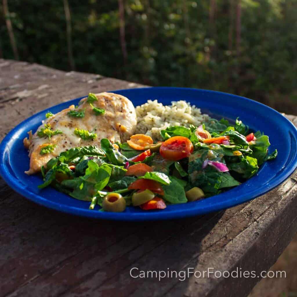 3 Ingredient Caesar Chicken by CampingForFoodies features a blue camping plate filled with a hearty dinner meal. The chicken breast is covered in a white, creamy sauce and golden brown Parmesan cheese. A bright green leafy salad with red cherry tomatoes that are sliced in half and purple onion are tossed in salad dressing that is next to a bed of white fluffy rice on the plate which is set on a wooden picnic table. A forest is in the background lit by the golden glow of a setting sun.