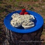 Camping Dutch Oven Biscuits And Gravy by CampingForFoodies features a blue camping plate filled with homemade Dutch oven biscuits topped with white creamy sausage gravy. Three bright red strawberries are set on the side of the plate that is set on top of a campfire log on a sunny morning.