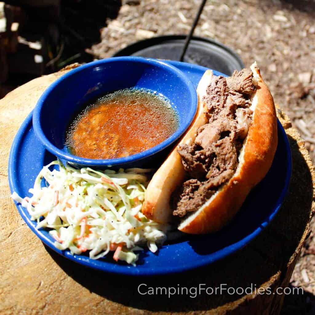 Dutch Oven French Dip Campfire Sandwiches by CampingForFoodies features a blue camping plate and bowl. A sliced beef sandwich in a hoagie roll sits on the side of the plate. Next to it, the bowl is filled with a brown au jus beef broth for dipping. A serving of coleslaw is on the side of the plate which is set on a campfire log with a black cast iron Dutch oven in the background on a sunny afternoon.