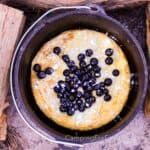 Lemon Blueberry Topped Campfire Dutch Baby by CampingForFoodies features a black cast iron Dutch oven filled with a golden brown, puffy Dutch baby pancake that is topped with fresh blueberries and sprinkled with white powdered sugar. The pot is set on the ground and surrounded with pieces of firewood that act as a wind block when baking in windy conditions.