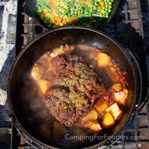 Campfire Pot Roast by CampingForFoodies features a black cast iron Dutch oven filled with a slow cooked beef roast surrounded by tender cubed potatoes in a reddish brown simmering liquid. The pot is set on a campfire grate next to a skillet filled with bright green, orange and yellow steamed green beans, peas, corn and carrots. The ash of campfire coals and charred firewood are in the background.