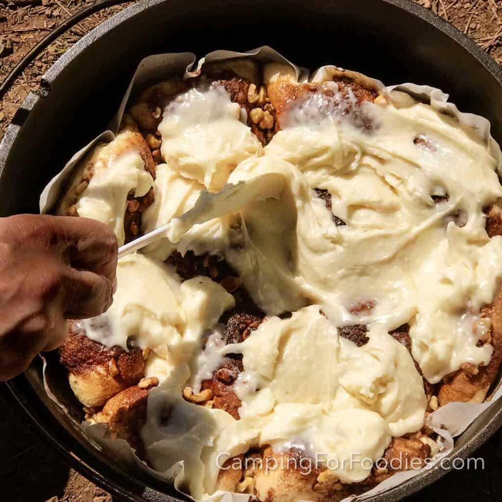 Camp Dutch Oven Cinnamon Rolls by CampingForFoodies features a black cast iron Dutch oven filled with golden brown cinnamon rolls that are topped with walnuts. A hand is holding a rubber spatula that is being used to spread cream cheese frosting over the rolls.