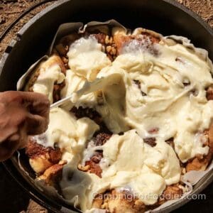 Camp Dutch Oven Cinnamon Rolls by CampingForFoodies features a black cast iron Dutch oven filled with golden brown cinnamon rolls that are topped with walnuts. A hand is holding a rubber spatula that is being used to spread cream cheese frosting over the rolls.