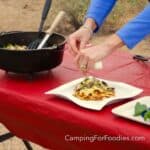 Dutch Oven Lasagna by CampingForFoodies features a black cast iron Dutch oven filled with homemade lasagna. The pot is set on a table covered in a red tablecloth. A single slice is set on a white plate, two hands are sprinkling freshly grated Parmesan cheese on the meal that is topped with ribbons of bright green fresh basil.