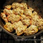 Sausage And Cream Cheese Stuffed Peppers by CampingForFoodies features a black cast iron Dutch oven set on a campfire grill grate. The pot is filled with red, yellow and orange mini bell peppers that are cut in half and stuffed with a creamy mixture of ground Italian pork sausage and cream cheese. The appetizers have been baked and are ready to be served.